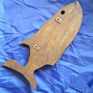 May include: Wooden whale-shaped wall decor. The whale is made of light brown wood with visible grain and features a tail, fins, and a hole near the top. Two metal brackets are attached for hanging. The decor is approximately 24 inches long.
