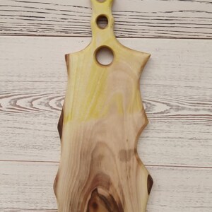 May include: A wooden serving board with a natural live edge and a light wood grain pattern. The handle features four circular cutouts of varying sizes arranged vertically, with a yellow-tinted finish applied to the upper handle section.