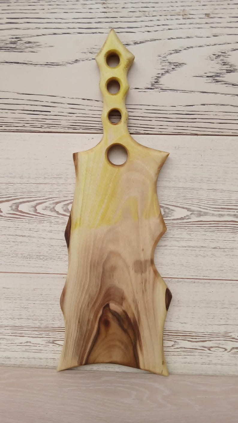 May include: A wooden serving board with a natural live edge and a light wood grain pattern. The handle features four circular cutouts of varying sizes arranged vertically, with a yellow-tinted finish applied to the upper handle section.