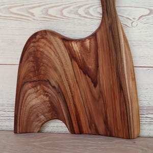 May include: A wooden serving board featuring a natural brown grain pattern with dark streaks. The board has an asymmetrical, curved shape with a distinct handle at the top that includes a circular hole for hanging, showcasing the wood's organic texture.