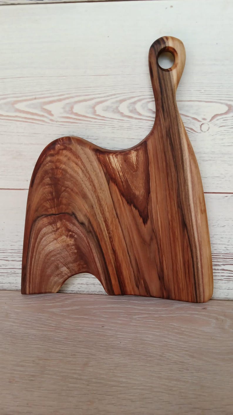 May include: A wooden serving board featuring a natural brown grain pattern with dark streaks. The board has an asymmetrical, curved shape with a distinct handle at the top that includes a circular hole for hanging, showcasing the wood's organic texture.