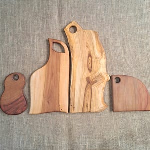 Puede incluir: Cuatro tablas de cortar de madera de diversas formas y tama&ntilde;os, cada una con un patr&oacute;n de veta natural y un orificio circular cerca de la parte superior para colgar. Las tablas muestran tonos marrones claros y oscuros con bordes org&aacute;nicos.