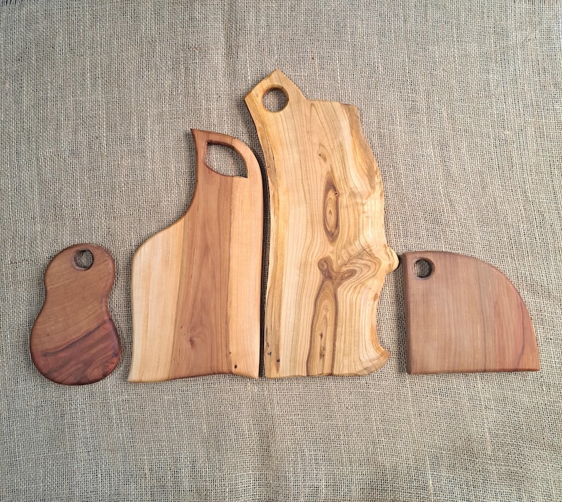 Puede incluir: Cuatro tablas de cortar de madera de diversas formas y tama&ntilde;os, cada una con un patr&oacute;n de veta natural y un orificio circular cerca de la parte superior para colgar. Las tablas muestran tonos marrones claros y oscuros con bordes org&aacute;nicos.