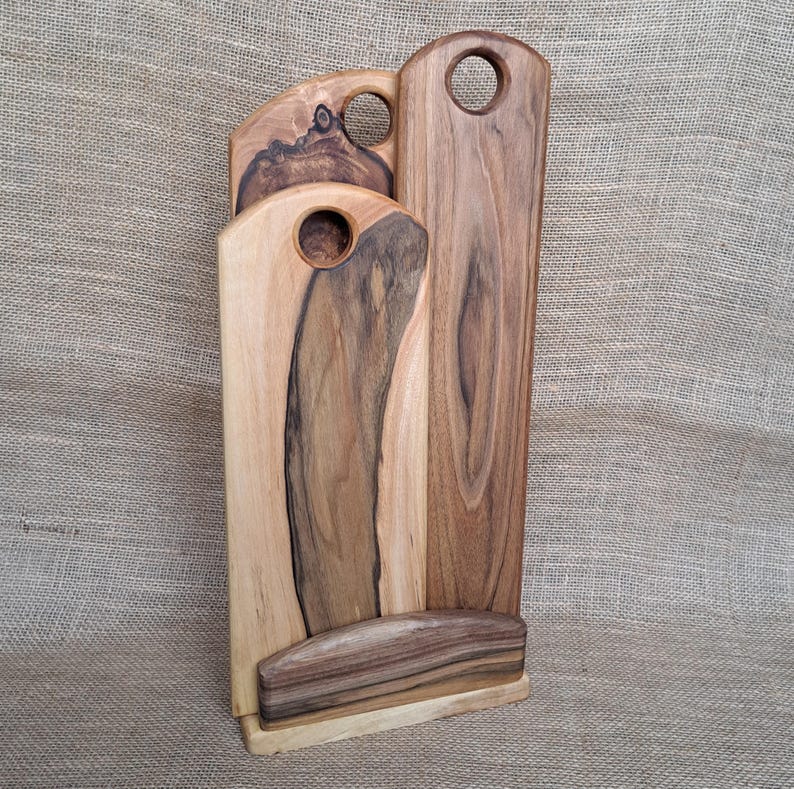 Op de afbeelding: Een set van drie verticale houten snijplanken met natuurlijke houtnerven in donkerbruine en lichtbruine tinten. Elke plank heeft een rond gat aan de bovenkant om op te hangen en ze staan samen in een houten standaard als basis.