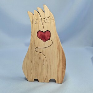 May include: Wooden cat ornament featuring two cats holding a red heart. The cats are crafted from light-coloured wood, with etched details for the eyes and whiskers. The heart is a darker red, providing contrast.