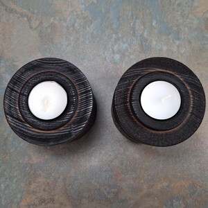 May include: Two circular, black wooden candle holders with a concentric design. Each holds a white tea light candle. The wood grain is visible, adding texture to the dark, rustic aesthetic. Suitable for home decor.