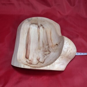 May include: A hand-carved wooden bowl with a natural wood grain finish. The bowl has a unique, organic shape with a rounded side and a more angular, open section. The wood's colour ranges from light tan to medium brown.