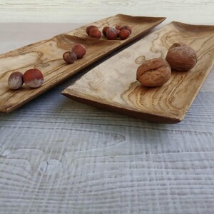 May include: Two rectangular wooden serving trays with a natural light brown wood grain pattern. The left tray contains several small brown hazelnuts, while the right tray holds two larger, textured brown walnuts on its smooth surface.