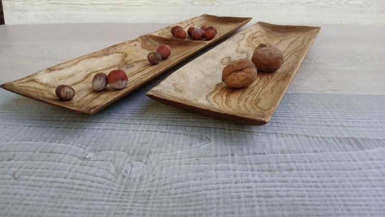 May include: Two rectangular wooden serving trays with a natural light brown wood grain pattern. The left tray contains several small brown hazelnuts, while the right tray holds two larger, textured brown walnuts on its smooth surface.