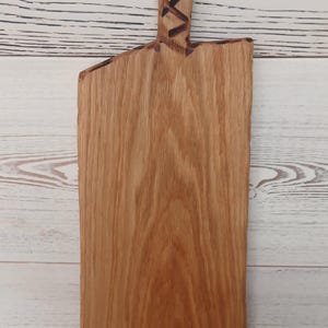 Pode incluir: Uma t&aacute;bua de servir retangular de madeira com borda natural e padr&atilde;o de gr&atilde;o vertical. O cabo apresenta um recorte circular e um design geom&eacute;trico de cruz queimado em tom escuro. A madeira &eacute; de cor castanho-claro quente com textura vis&iacute;vel.