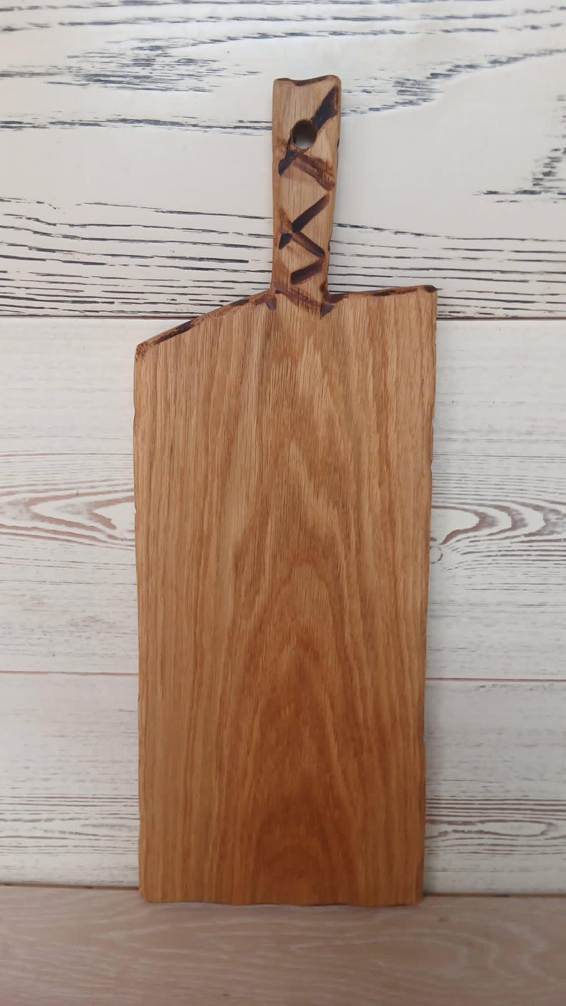Pode incluir: Uma t&aacute;bua de servir retangular de madeira com borda natural e padr&atilde;o de gr&atilde;o vertical. O cabo apresenta um recorte circular e um design geom&eacute;trico de cruz queimado em tom escuro. A madeira &eacute; de cor castanho-claro quente com textura vis&iacute;vel.