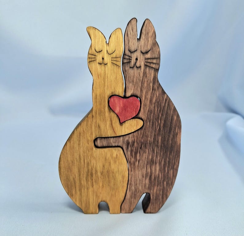 以下が含まれることがあります： 木製の猫の置物。目を閉じた2匹の猫が抱き合っています。1匹は薄茶色、もう1匹は濃い茶色で、赤いハートを持っています。魅力的で手作りの装飾品です。