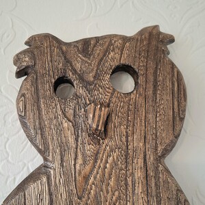 May include: A wooden owl carving with a detailed grain pattern. The owl has large, round eye holes and a carved beak. The wood is a warm brown color, and the carving has a rustic, handcrafted appearance.