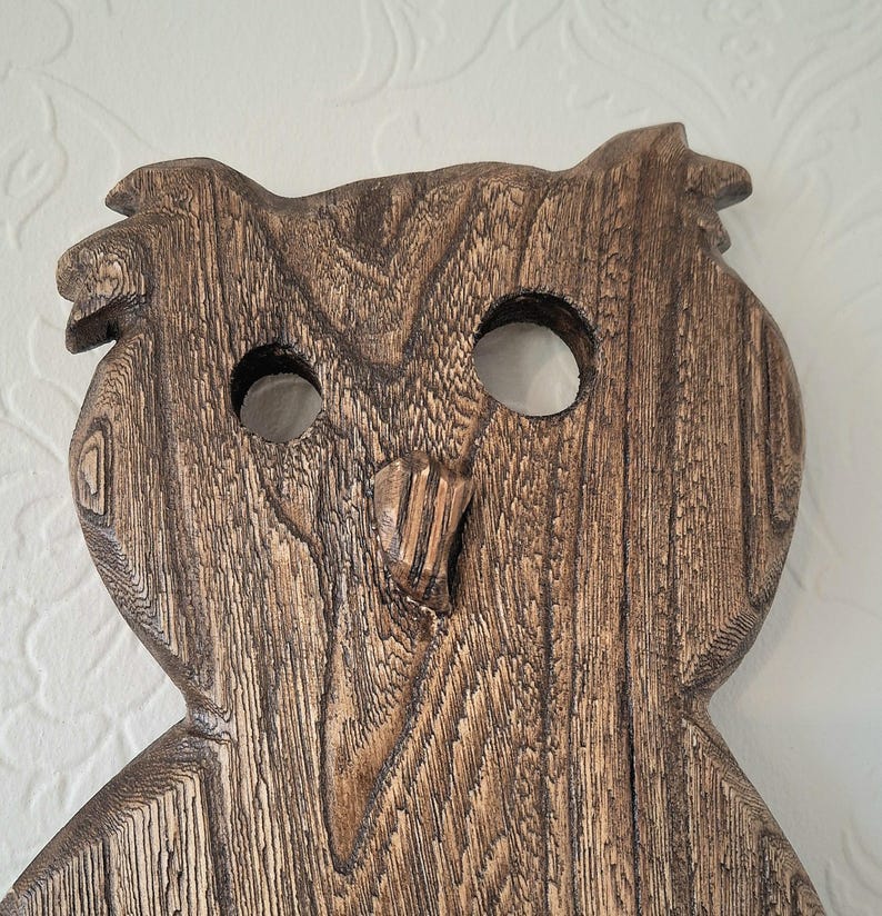 May include: A wooden owl carving with a detailed grain pattern. The owl has large, round eye holes and a carved beak. The wood is a warm brown color, and the carving has a rustic, handcrafted appearance.