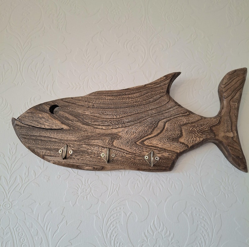 May include: A wooden fish-shaped key holder with a dark brown finish. The key holder has a carved design and features three metal hooks for hanging keys. The fish is mounted on a white wall.