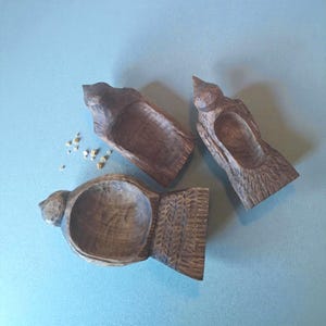 May include: Three hand-carved wooden bird-shaped bowls. The bowls are brown with a textured finish, and each has a hollowed-out interior. Small, light-colored granules are scattered near one bowl. The bowls are arranged on a light blue surface.