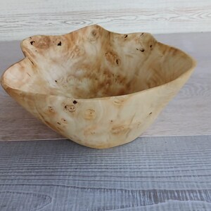 Op de afbeelding: Een handgemaakte houten kom met een unieke, organische vorm. De kom is gemaakt van lichtgekleurd hout met een natuurlijke nerf en knoestenpatroon. De rand heeft een golvende, onregelmatige rand, wat bijdraagt aan de rustieke charme.