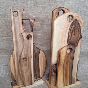 Op de afbeelding: Twee houten standaards met meerdere snijplanken van natuurlijk hout in verschillende vormen en maten. Elke plank heeft een uniek nerfpatroon met licht- en donkerbruine tinten en een cirkelvormige uitsparing aan de bovenkant als handvat.