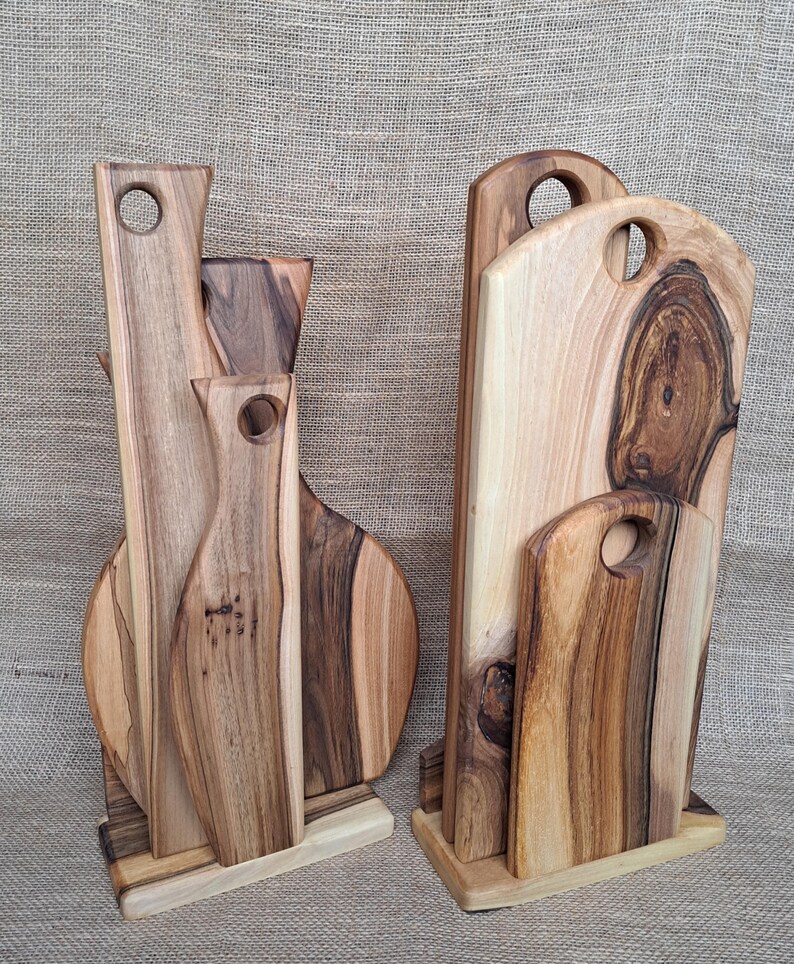 Op de afbeelding: Twee houten standaards met meerdere snijplanken van natuurlijk hout in verschillende vormen en maten. Elke plank heeft een uniek nerfpatroon met licht- en donkerbruine tinten en een cirkelvormige uitsparing aan de bovenkant als handvat.
