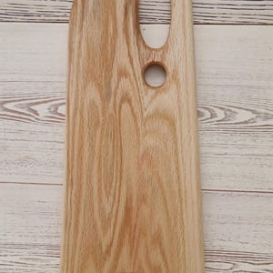 May include: A light brown wooden serving board with a natural wood grain pattern. The board features an elongated vertical shape with a curved top edge, a large oval cutout handle, and a smaller circular hole below it, ending in a concave base.