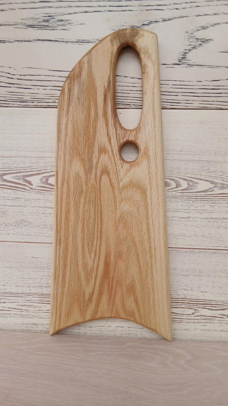 May include: A light brown wooden serving board with a natural wood grain pattern. The board features an elongated vertical shape with a curved top edge, a large oval cutout handle, and a smaller circular hole below it, ending in a concave base.