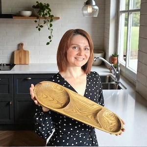 Könnte beinhalten: Ein Holzserviertablett mit drei geschnitzten Abschnitten. Das Tablett hat eine hellbraune Farbe mit einem Holzmaserungsmuster. Das Tablett wird vor einem Küchenhintergrund gehalten.