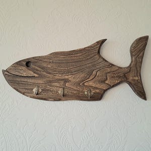 May include: A wooden fish-shaped wall hook with a dark brown finish. The fish has a carved design and three metal hooks along its belly. The eye is a small, round hole. The tail and fins are also carved.