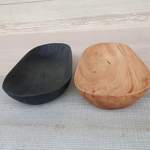 May include: Two wooden bowls, one black and one natural wood, sit side-by-side. The black bowl has a smooth, matte finish. The natural wood bowl displays the grain and knots of the wood. Both bowls have an organic, asymmetrical shape.