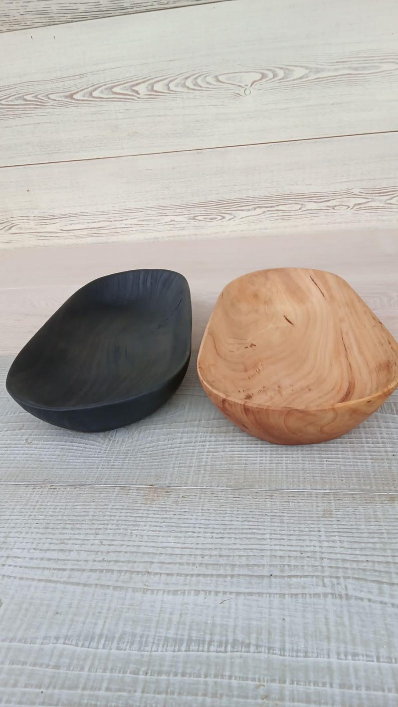 May include: Two wooden bowls, one black and one natural wood, sit side-by-side. The black bowl has a smooth, matte finish. The natural wood bowl displays the grain and knots of the wood. Both bowls have an organic, asymmetrical shape.