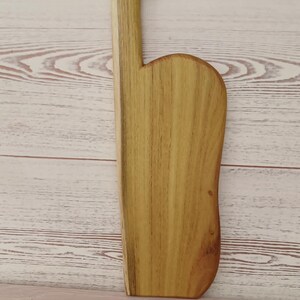 May include: A light brown wooden serving board with a natural wood grain pattern. The board features a long, straight side and a curved side, with a narrow handle at the top that includes a circular hole for hanging. The wood has a smooth finish.