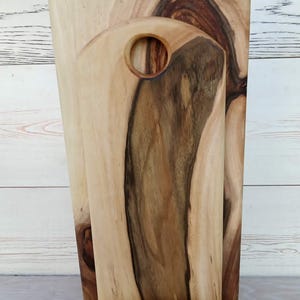 Op de afbeelding: Een set van twee houten snijplanken met natuurlijke houtnerven, elk voorzien van een rond handvatgat aan de bovenkant. De planken staan rechtop in een bijpassende houten voet en vertonen licht- en donkerbruine houttinten met zichtbare noesten.