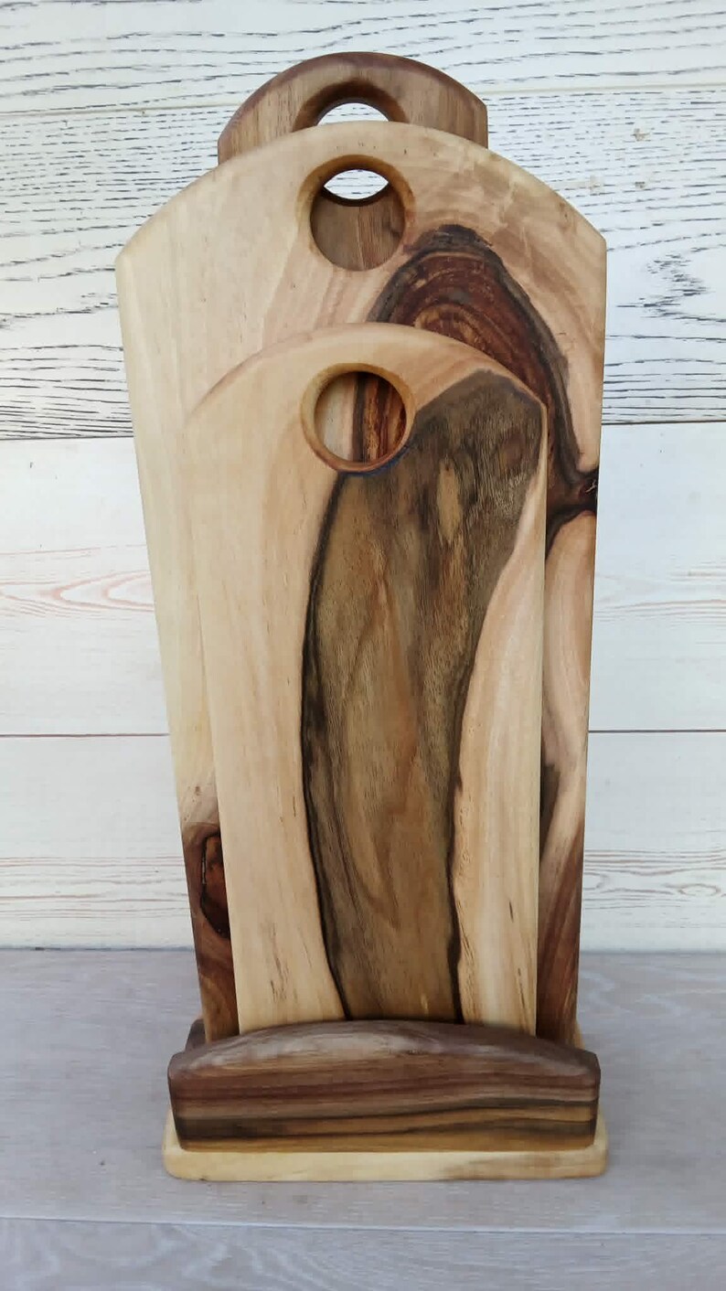 Op de afbeelding: Een set van twee houten snijplanken met natuurlijke houtnerven, elk voorzien van een rond handvatgat aan de bovenkant. De planken staan rechtop in een bijpassende houten voet en vertonen licht- en donkerbruine houttinten met zichtbare noesten.