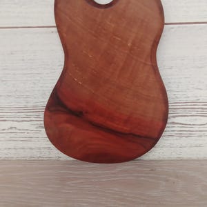 Puede incluir: Una tabla de servir de madera de color marr&oacute;n rojizo, con una forma org&aacute;nica irregular y un orificio circular cerca de la parte superior para colgarla. La veta de la madera es visible en toda la superficie, mostrando variaciones naturales.