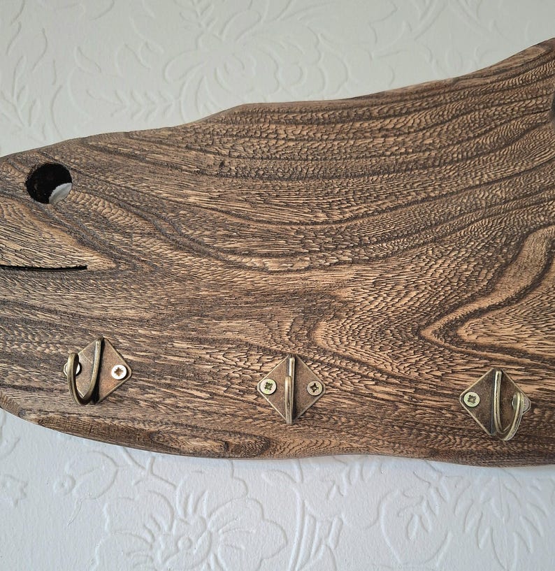 May include: A rustic wooden wall-mounted key holder with three metal hooks. The wood has a dark brown finish with visible grain patterns. The hooks are diamond-shaped with a bronze finish, perfect for organizing keys or small items.