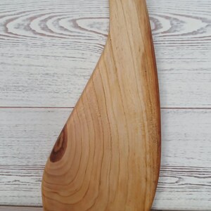 May include: A light brown wooden cutting board with a curved, asymmetrical shape and a smooth, polished finish. The board features a prominent natural wood grain pattern, a visible knot near the base, and a circular hole at the top for hanging.