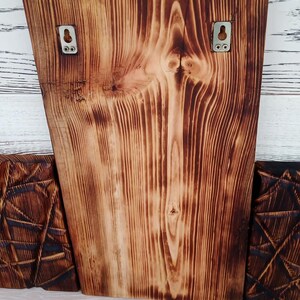 May include: Wooden wall art with a natural wood grain and a burnt finish. Two metal hangers are attached to the back. The piece is flanked by two other wooden panels with geometric designs. The wood grain is visible.