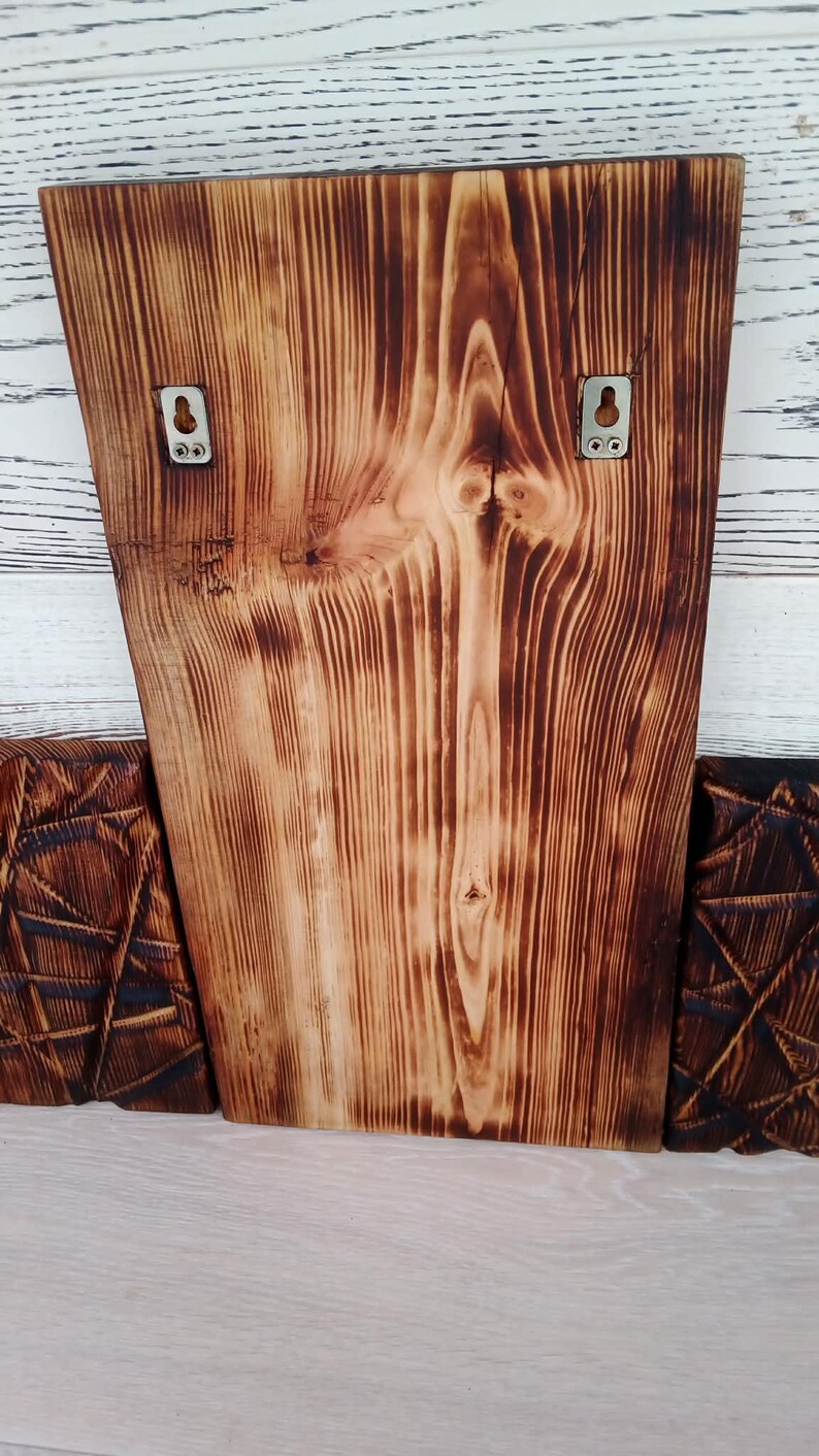 May include: Wooden wall art with a natural wood grain and a burnt finish. Two metal hangers are attached to the back. The piece is flanked by two other wooden panels with geometric designs. The wood grain is visible.