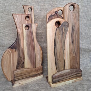 Op de afbeelding: Twee sets houten snijplanken met natuurlijke houtnerven, elk bestaande uit drie planken van verschillende hoogtes die rechtop worden gehouden door een bijpassende houten voet. Elke plank heeft een rond gat aan de bovenkant om op te hangen.