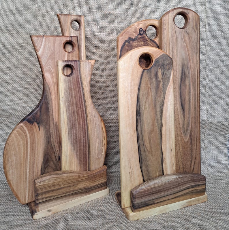 Op de afbeelding: Twee sets houten snijplanken met natuurlijke houtnerven, elk bestaande uit drie planken van verschillende hoogtes die rechtop worden gehouden door een bijpassende houten voet. Elke plank heeft een rond gat aan de bovenkant om op te hangen.