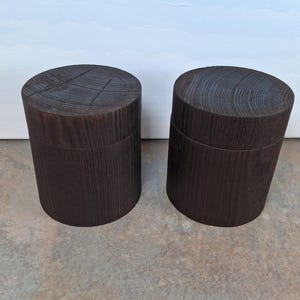 May include: Two dark brown wooden containers. Each has a cylindrical shape and a lid. The wood grain is visible on the top and sides. The containers are placed side by side on a neutral surface.