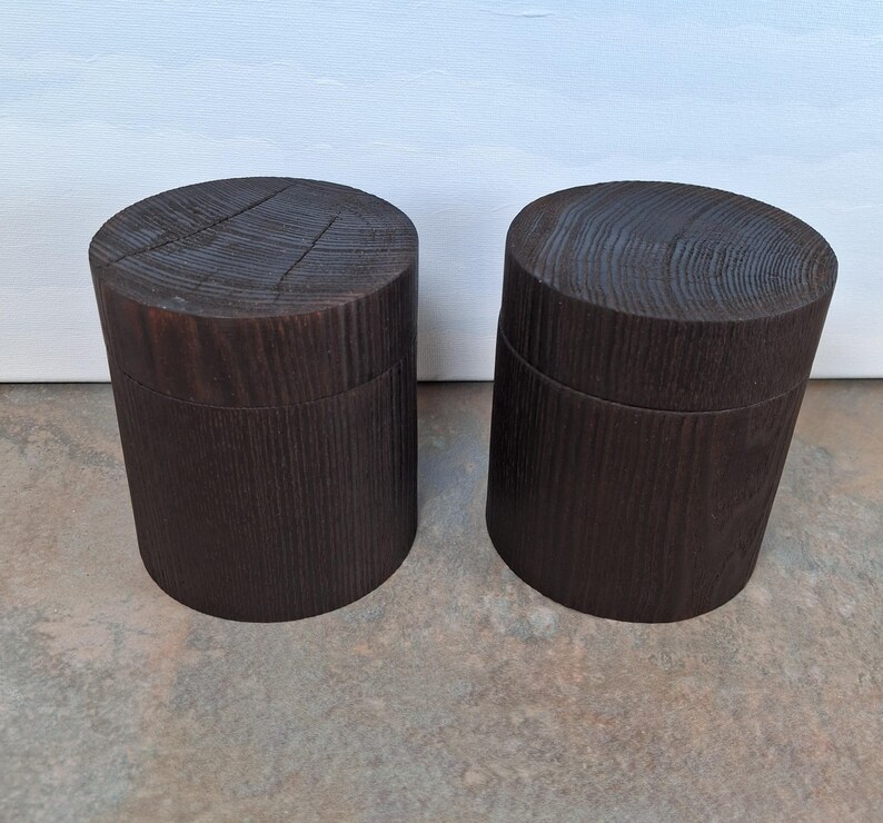 May include: Two dark brown wooden containers. Each has a cylindrical shape and a lid. The wood grain is visible on the top and sides. The containers are placed side by side on a neutral surface.