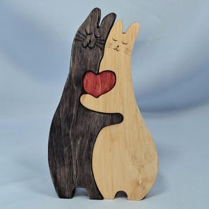 以下が含まれることがあります： 木製の猫の置物。濃い茶色と薄い茶色の2匹の猫が抱き合い、赤いハートを持っています。猫は目を閉じ、ひげがあり、愛情深く気まぐれなデザインです。