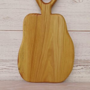 May include: A light brown wooden cutting board with a smooth, polished finish and a distinct pear-shaped silhouette. The board features a prominent handle at the top with a circular cutout, showcasing natural wood grain patterns throughout the surface.