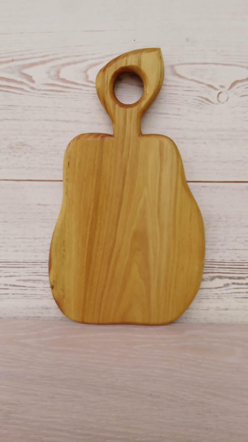 May include: A light brown wooden cutting board with a smooth, polished finish and a distinct pear-shaped silhouette. The board features a prominent handle at the top with a circular cutout, showcasing natural wood grain patterns throughout the surface.