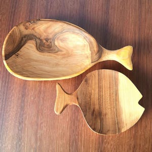 May include: Two wooden fish-shaped serving dishes. The larger dish is a bowl with a tail fin handle. The smaller dish is a flat serving board. Both are made of light and dark brown wood with visible grain.