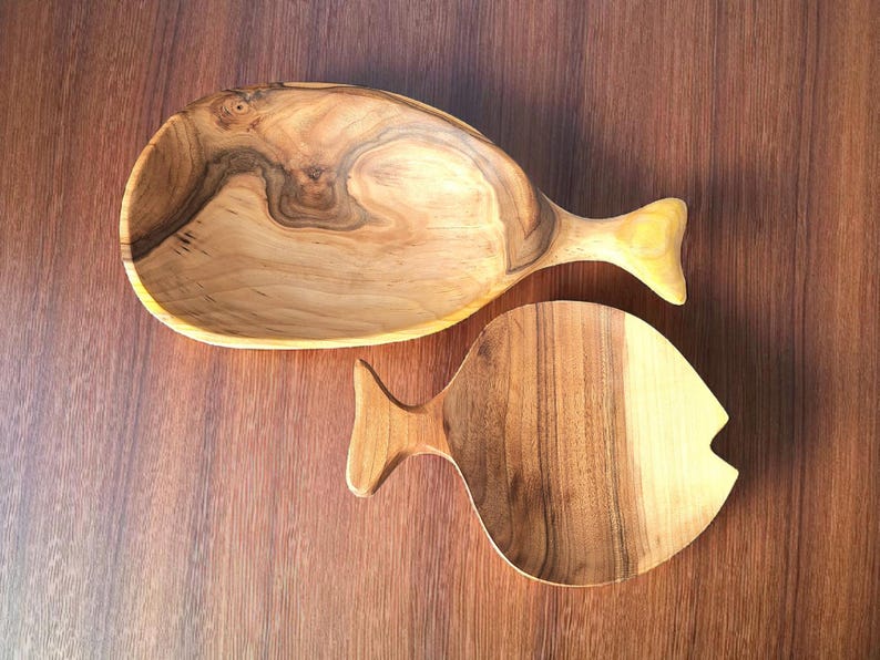 May include: Two wooden fish-shaped serving dishes. The larger dish is a bowl with a tail fin handle. The smaller dish is a flat serving board. Both are made of light and dark brown wood with visible grain.