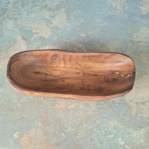 May include: A handcrafted wooden bowl with a natural, organic shape. The bowl is made from a single piece of wood, showcasing the wood grain and knots. The bowl's interior is smooth, while the exterior has a slightly rough texture. The bowl is approximately 12 inches long.