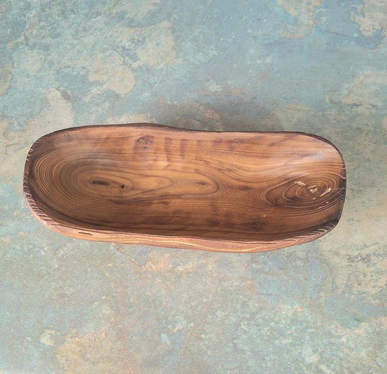 May include: A handcrafted wooden bowl with a natural, organic shape. The bowl is made from a single piece of wood, showcasing the wood grain and knots. The bowl's interior is smooth, while the exterior has a slightly rough texture. The bowl is approximately 12 inches long.