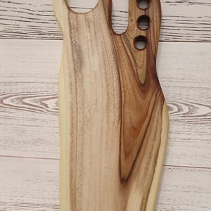 May include: A wooden serving board with a natural live edge, featuring a large circular handle opening at the top left and a vertical row of four smaller circular cutouts along the right side, showcasing a light and dark brown wood grain pattern.