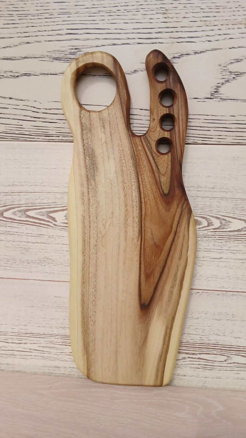 May include: A wooden serving board with a natural live edge, featuring a large circular handle opening at the top left and a vertical row of four smaller circular cutouts along the right side, showcasing a light and dark brown wood grain pattern.